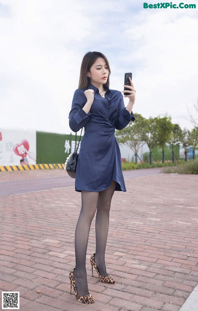 A woman in a blue shirt dress is taking a selfie.