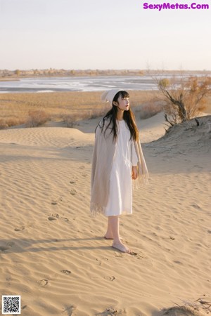 A woman in a white dress and hat walking on a sandy beach.