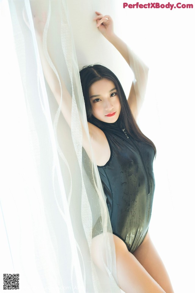 A woman in a black bodysuit leaning against a white curtain.